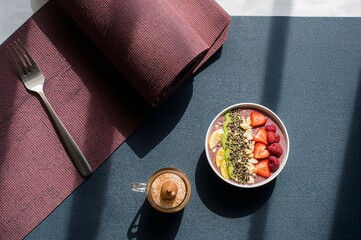 Overhead view of a smoothie bowl with fruit, a fork, and a drink on a textured surface in sunlight