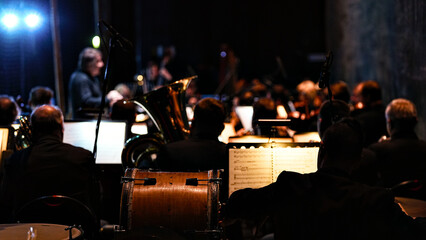 Close up of classical orchestra performance featuring cello instrument in foreground with blurred musicians and sheet music under warm stage lighting