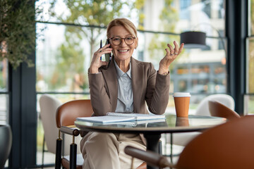 Mature businesswoman smiling, talking on smartphone in cafe