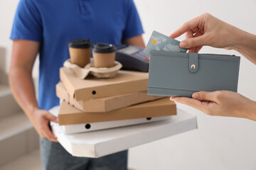 Female hands taking credit card out of wallet near courier with pizza boxes and coffee in stairway, closeup