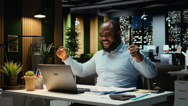 Overjoyed delighted guy reading good news in his emails at night, celebrating a career breakthrough and beaming in the office. Businessman with awesome opportunity and contract offer.