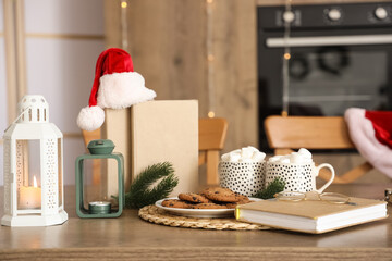 Books with Santa hat, Christmas lanterns, cookies and cocoa on table in kitchen