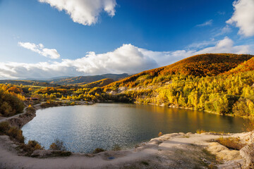 Obraz premium A lake nestled between autumn hills, with golden trees reflecting in the water under a bright blue sky and scattered clouds. Flooded quarry near the village Sutovo in Slovakia, Europe.