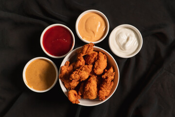 Top view of a bowl of fried chicken with different sauces on top of a black cloth.