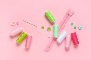 Colorful thread spools with buttons, zipper and needle on pink background