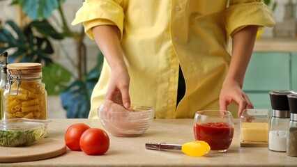 A woman in a yellow shirt arranging fresh ingredients, including tomatoes, chicken, and pasta, on a kitchen countertop for cooking.