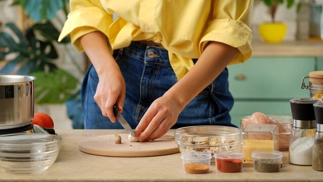 An unrecognizable woman in jeans and a yellow shirt, cutting mushrooms on a cutting board, surrounded by spices and ingredients. Preparing lunch or dinner in a home kitchen. - Powered by Adobe