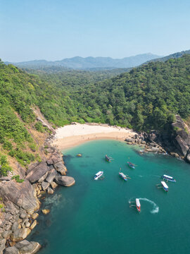 A stunning aerial top view of Butterfly Beach in Goa, showcasing golden sands waters and lush green hills in the backdrop. People are seen enjoying the beach while a boat sails nearby.