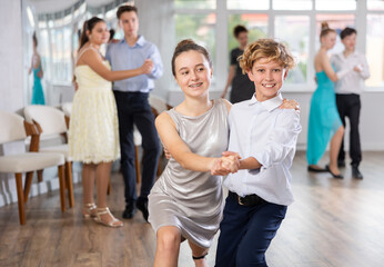 Obraz premium Cheerful teenage boy and girl confidently performing tango step at choreography lesson, surrounded by fellow students learning dance