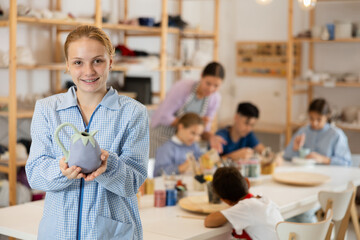 Joyful teenage girl holds a clay product against the background of kids and a teacher. Children learn with interest at the ceramics master class