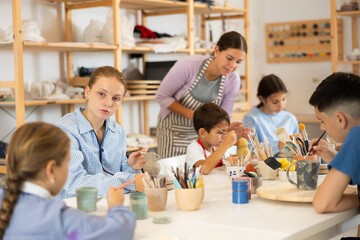 Children paint ceramics with brushes at the table in the creative studio. Teacher shows and helps children at the master class on ceramics