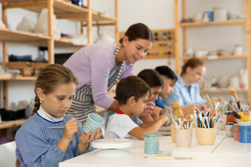 Interested little girl painting handmade ceramic cup with brush in pottery workshop