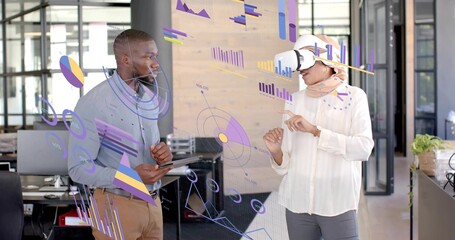 Presenting data by two colleagues in office, one wearing hijab with VR headset, tablet and plant