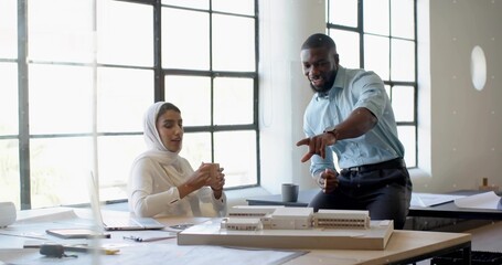 Collaborating architects discussing scale model at design studio table, with blueprints and laptop