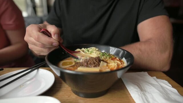 Close-up of man in black shirt dining on ramen with meat and egg slices Man eating ramen soup with chopsticks at table in restaurant, dark bowl and egg visible