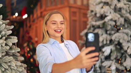 Happy blonde woman takes Christmas selfie video on smartphone. Joyful emotions radiate from smiling face, celebrating holiday season. Communication and digital connection captured video chat online - Powered by Adobe