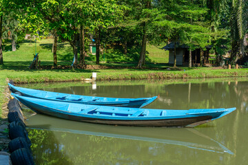 Two blue wooden boats are moored on the water's surface, with trees and a small wooden hut in the background. The scene is illuminated by bright sunlight, creating reflections on the water's surface.