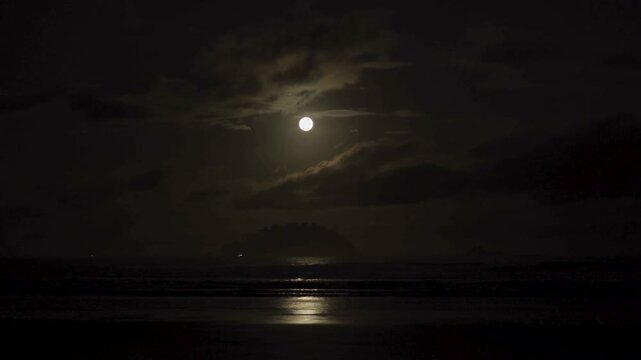A bright full moon casts silver reflections across the dark ocean as soft clouds drift through a deep blue night sky, creating a peaceful and mysterious seascape.