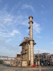 Tall industrial exhaust stack and complex machinery rising against a bright blue sky with scattered white clouds