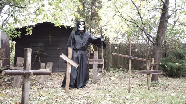 Grim reaper or Death with a skeleton head in an old cemetery graveyard holding a scythe with wood crosses in the rain. Gloomy Halloween horror. Slider shot in slow motion.