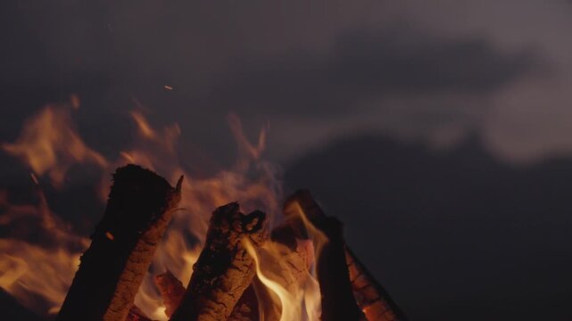 Bright campfire burning in the foreground with a mountain range and lake in the background at night. Peaceful outdoor scene symbolizing warmth, adventure, and solitude in nature.
