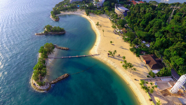 Aerial view of Palawan Beach, Sentosa, Singapore, featuring turquoise water, white sand, palm trees, and a small connected island
