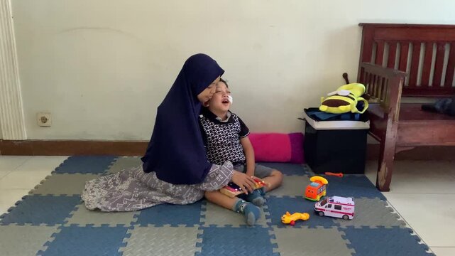 A mother wearing a hijab lovingly hugs her crying child surrounded by toys on a play mat. Capturing moments of deep emotional comfort and resilience.