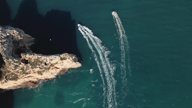 Aerial drone steadily moves forward parallel to a boat and group of kayaks, both cutting smooth wakes across the turquoise waters and casting dramatic shadows near the sheer cliffs of Ponta da Piedade