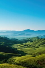 Fototapeta premium Rolling green hills meet misty mountains under clear blue sky