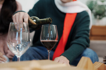 Couple wearing Santa hats celebrating Christmas at home, smiling while pouring red wine, surrounded by gifts and festive decorations.
