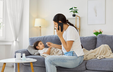Fototapeta premium Mother checking temperature of child while calling doctor for consultation about daughters cold. Sick tired child with fever and flu lying under blanket on living room sofa, parent holding thermometer