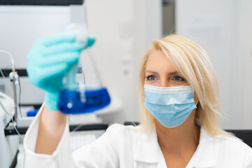 Middle aged female chemist wearing mask and lab coat observing blue liquid in flask in laboratory.