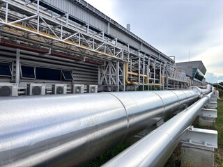 Long, large industrial stainless steel pipelines run horizontally outside a modern factory building under an overcast sky. The piping is part of a complex process system with smaller pipes and support