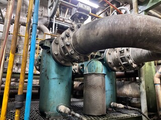 Heavy-duty industrial piping with large flanges and a strainer sitting on a metal grate floor inside a power station or factory. The turquoise and gray colors highlight old, complex machinery and infr