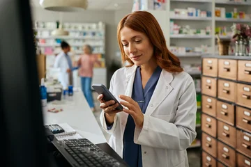 Fotobehang Beren Smiling female pharmacist using cell phone while working in pharmacy.  © Drazen