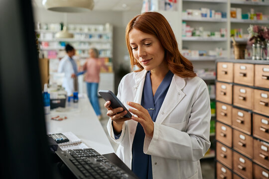 Smiling female pharmacist using cell phone while working in pharmacy. - Powered by Adobe