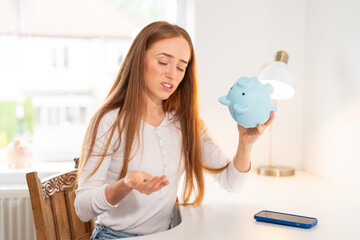 Young woman looking disappointed while checking her savings in a blue piggy bank at home during the day