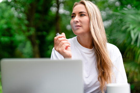 Woman enjoying a moment outdoors while vaping near a laptop in a lush green environment