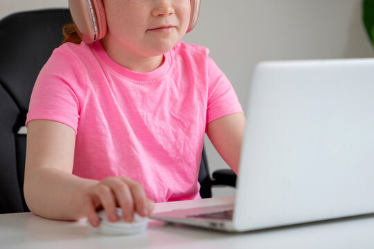 Child in pink shirt using laptop with headphones while learning at home during the day