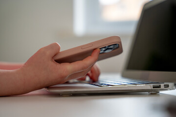 Person using a smartphone while typing on a laptop at a workstation in a bright and modern office environment