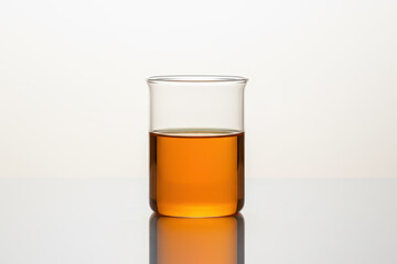 Amber liquid in clear glass beaker on reflective surface with white background, showcasing laboratory or cosmetic oil sample in minimal studio lighting