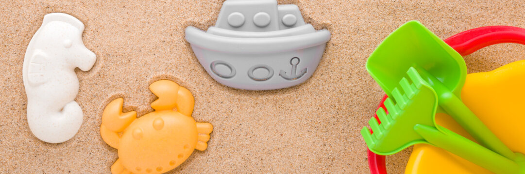 Colorful different plastic shapes and bucket with spade and rake on light brown sea sand background. Closeup. Playing toys for baby. Flat lay. Wide banner. Top down view. - Powered by Adobe