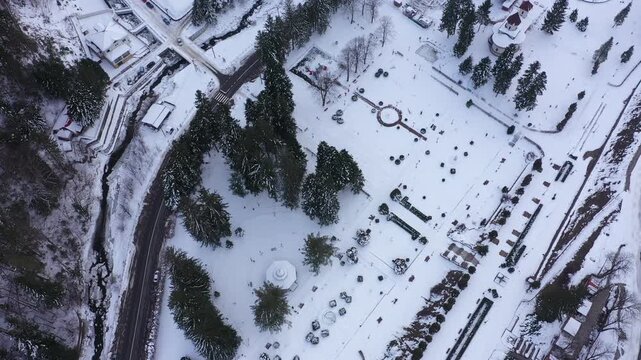 Ascending aerial view, drone, of Slanic Moldova's central park, Romania, covered in white snow. A road, river, and buildings surround the idyllic winter landscape.