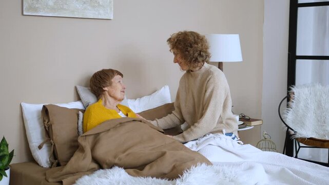 Kind adult daughter taking care of her sick senior mother lying in bed at home, showing love and support by holding her hand and talking with her, representing family relationships and caregiving