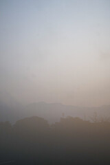 ​A soft, hazy morning or evening landscape showing silhouettes of hills and trees covered in a thick layer of fog or smog against a pale, muted sky.