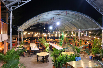A spacious outdoor restaurant at night with a large covered awning, featuring many wooden tables and chairs, lush potted plants, and strung warm yellow fairy lights.