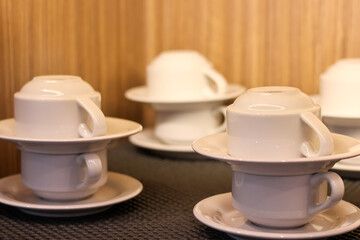 Neatly stacked white ceramic coffee or tea cups and saucers, arranged for self-service on a textured mat with a blurred light-brown wooden background.