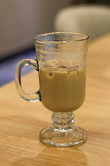 Close-up of a half-drunk glass mug of iced coffee with milk, featuring condensation on the glass and resting on a light wooden table in a dimly lit setting.