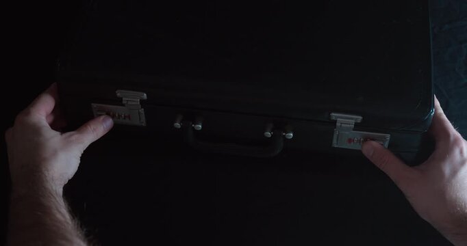 A person unlocks a closed black briefcase under moody lighting without opening it. Ideal for thriller, mystery, espionage, or crime-related visual storytelling.