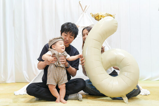 A man and a woman are holding a baby and a balloon that says six. The baby is smiling and the balloon is white. The scene is happy and joyful, as the family is celebrating the baby's first birthday